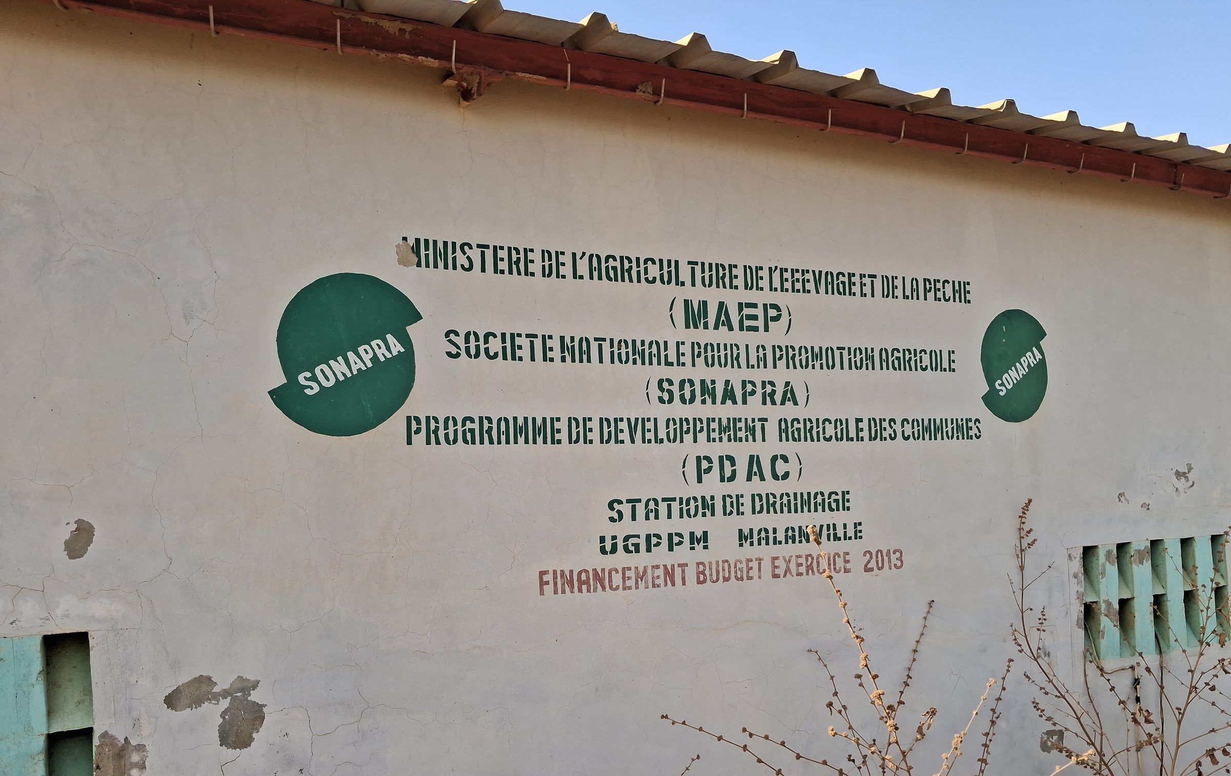 Mur blanc écaillé portant des logos verts et le texte « Station de drainage UGPPM Malanville », bouches d'aération à droite.
