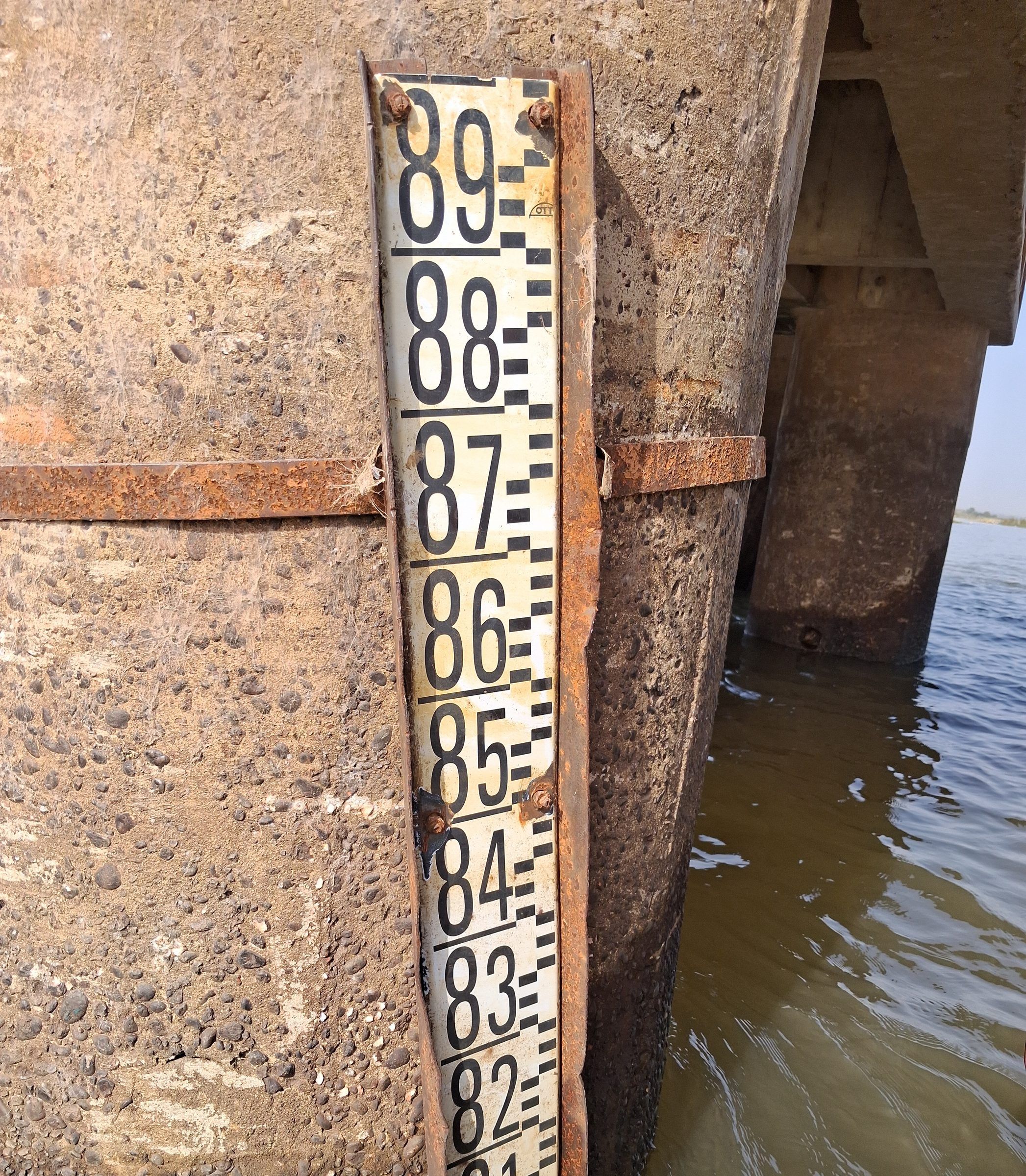 Règle graduée noire et blanche, numérotée de 82 à 89, sanglée contre une pile en béton qui plonge dans l'eau.