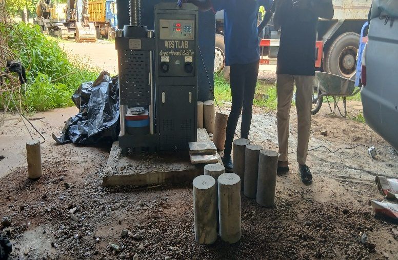 Presse d'essai installée en plein air, éprouvettes cylindriques alignées à côté ; plusieurs personnes autour.