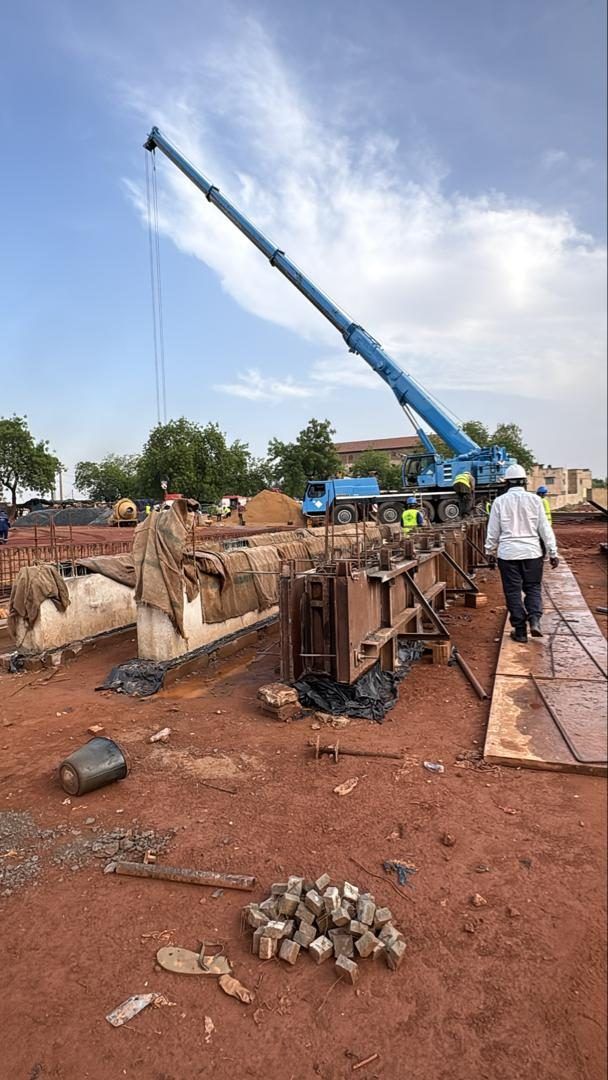 Flèche bleue d'un camion-grue dressée vers le ciel au-dessus des coffrages ; un homme en blanc marche sur le chantier.